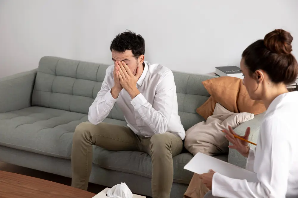 A person sits on a couch during a therapy session, covering their face with their hands while appearing overwhelmed and distressed. A mental health professional sits nearby, listening and taking notes, illustrating the emotional challenges associated with anxiety disorder and the process of seeking psychological support.