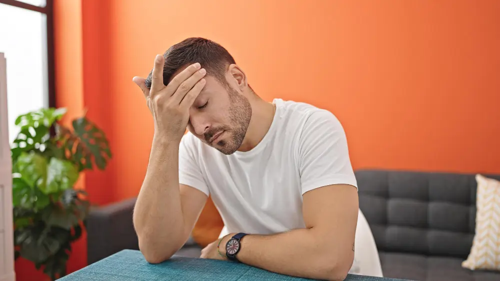 Man sitting indoors with his hand on his forehead, appearing stressed, fatigued, or overwhelmed, illustrating the effects of cortisol, chronic stress, anxiety, or mental health challenges.