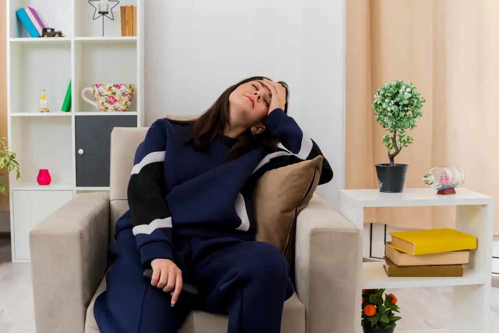 Exhausted woman slumped in armchair holding forehead, showing signs of adrenal fatigue and emotional burnout at home setting.
