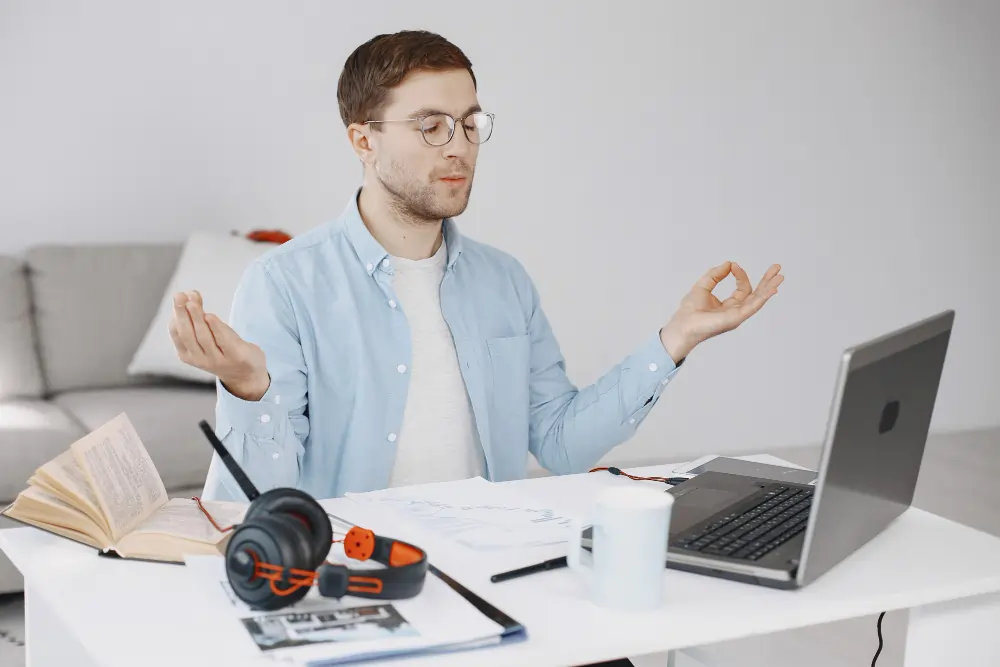 A person meditating while working at a desk with a laptop, books, and headphones, representing everyday stress management through practical, evidence-based strategies such as mindfulness, relaxation, and healthy work habits.