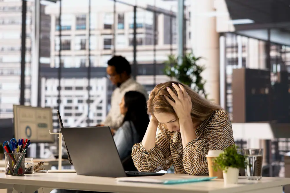 Overwhelmed office worker gripping her head beside laptop, surrounded by busy workplace, showing intense job burnout