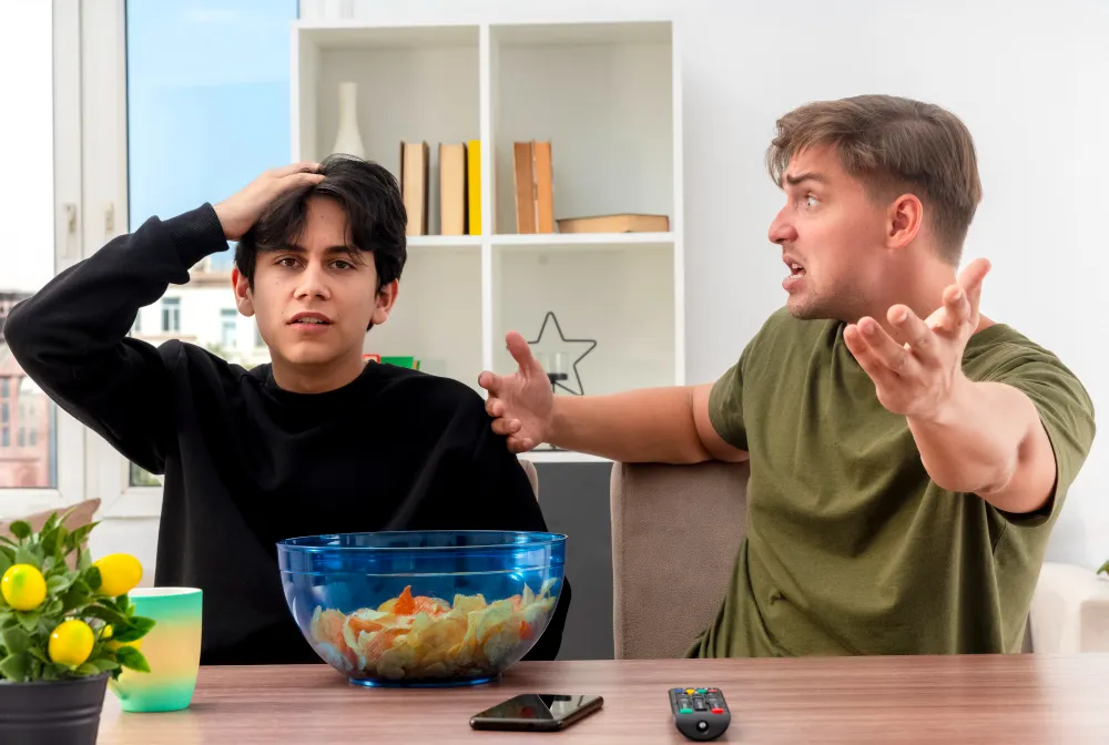 Frustrated man gesturing while arguing with a stressed young man sitting at a table with snacks and a remote control.
