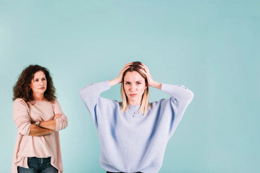 Overwhelmed young woman holds her head as an older woman watches with crossed arms, showing how ADHD in women is missed