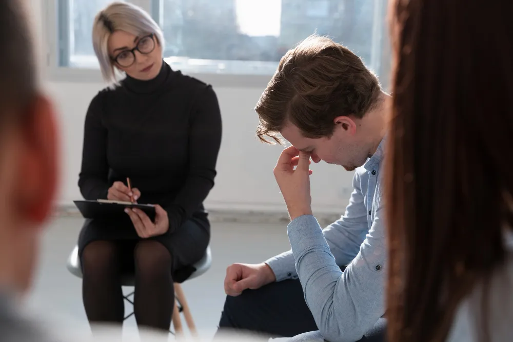 Woman taking notes while a man sits with his head down in a group therapy setting, illustrating the DSM-5 ADHD diagnostic criteria explained