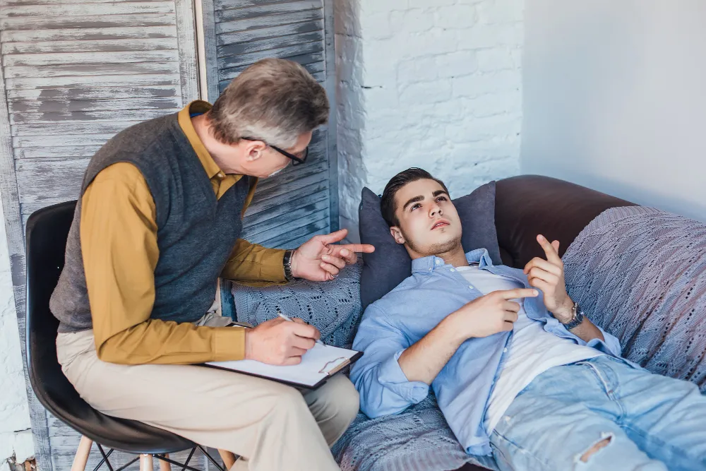 Male therapist taking notes while young adult patient reclines on couch during ADHD consultation